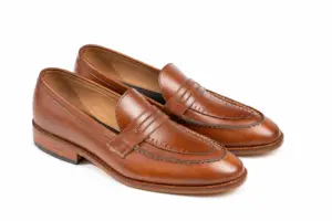 Three-quarter view of brown pull-up leather penny loafers showing cut-out penny strap, perforated apron border and brown leather grain