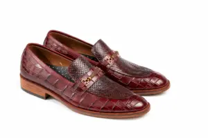 Three-quarter view of burgundy reptile-pattern horsebit loafers showing gold-tone bit-style hardware, lizard-pattern vamp and larger croc-pattern scales