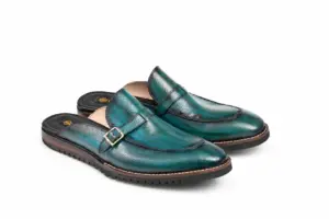 Three-quarter view of teal patina buckle mules with Vibram soles showing silver-tone buckle, teal patina, apron seam and light sole-edge stitching