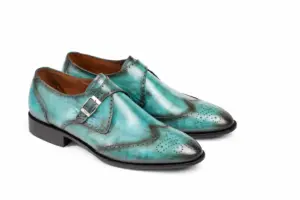 Three-quarter view of turquoise patina wingtip single monk strap shoes showing single silver-tone buckle, wingtip broguing and turquoise tonal patina